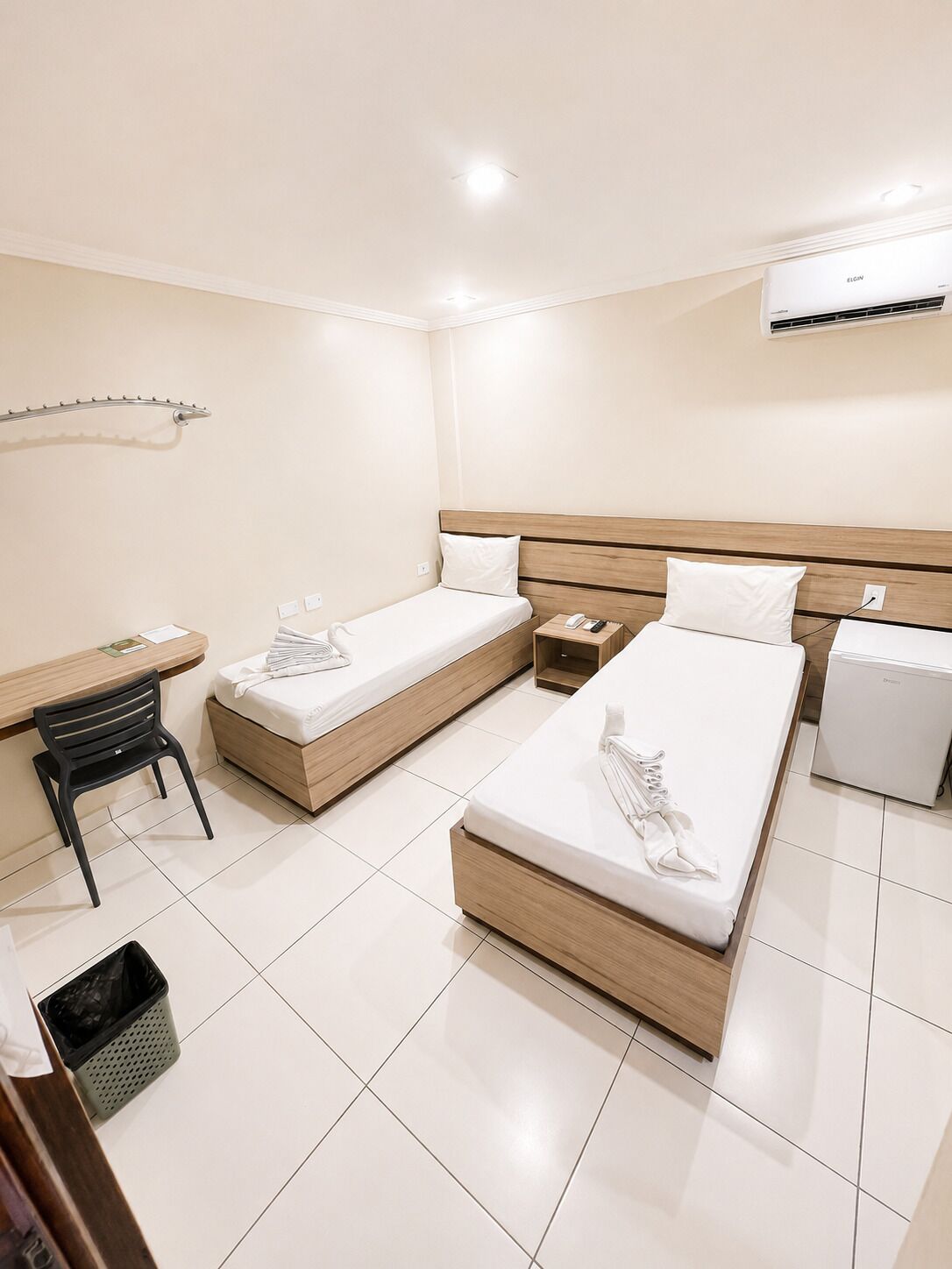 A hotel room with two single beds, a desk with a chair, a small bedside table, a trash bin, and an air conditioning unit.