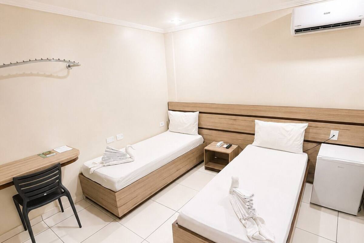 A hotel room with two single beds, a desk with a chair, a small bedside table, a trash bin, and an air conditioning unit.