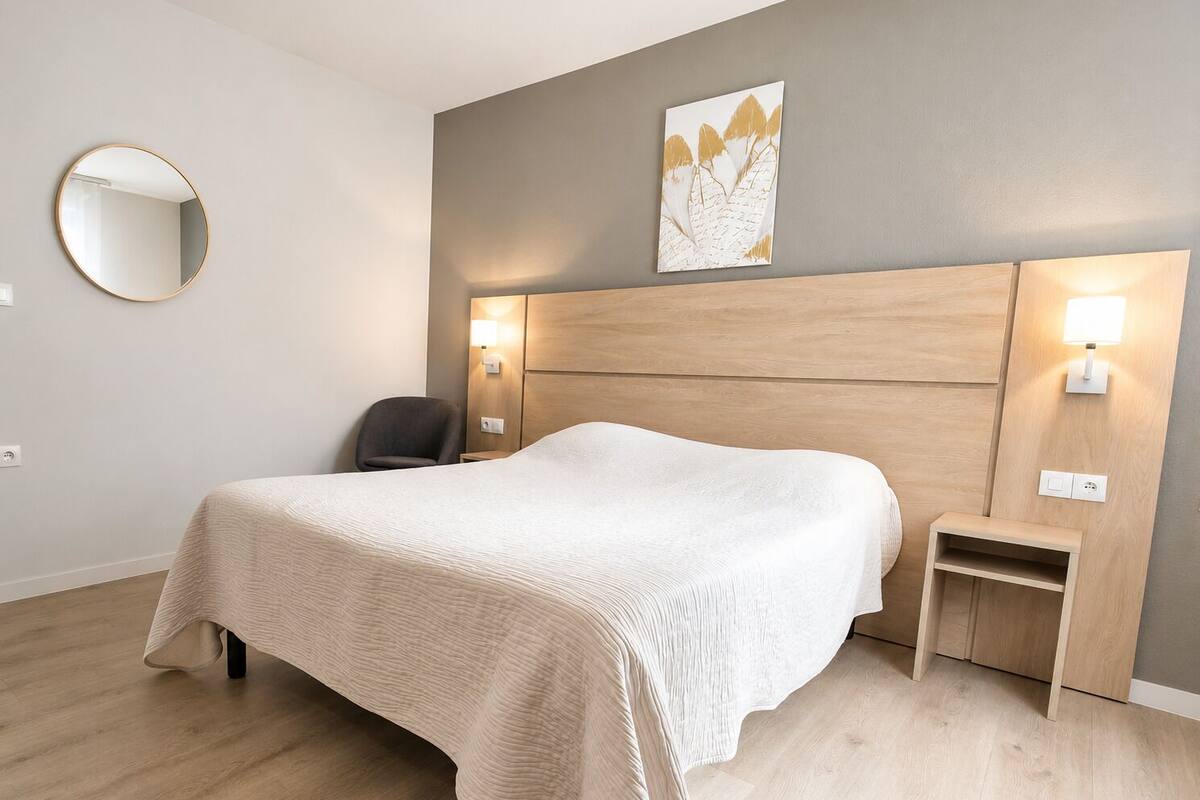Habitación de hotel con cama, mesita de noche, silla, lámpara de pared y un espejo redondo.