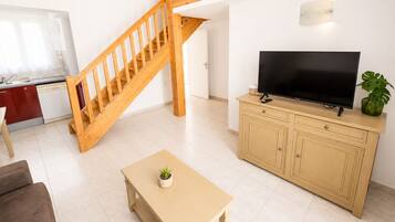 Een woonkamer met een houten trap, een televisie op een kast, een kleine tafel met een plant, en een keuken met een rode kast en witte apparaten.
