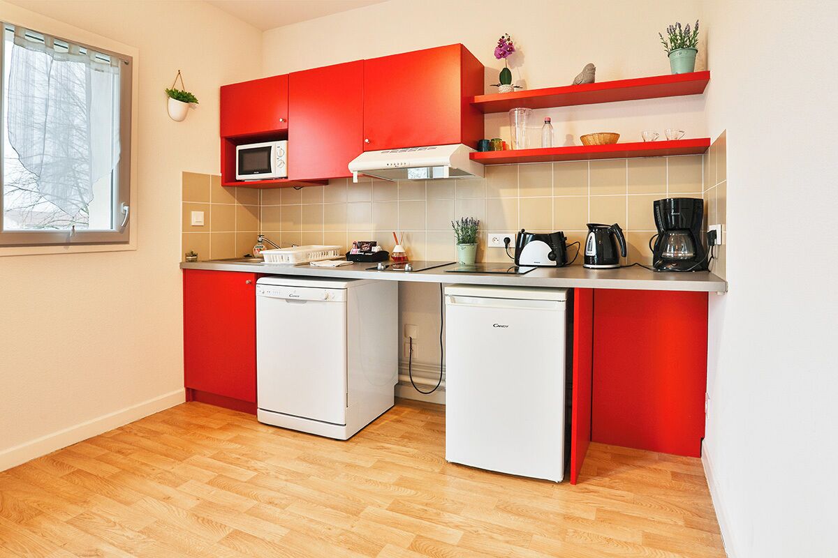 Une cuisine de taille réduite avec des meubles de rangement rouges, un four à micro-ondes, une machine à café et un lave-vaisselle.