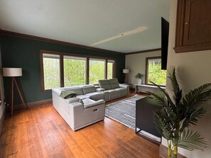 Un salon comprenant un canapé d’angle, une lampe sur pied, un radiateur et de grandes fenêtres donnant sur la forêt.