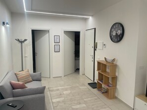 Ein modernes Wohnzimmer mit einem grauen Sofa, einem Holzregal, einer Uhr und einer Tür zu einem anderen Raum.