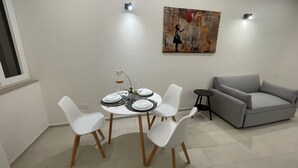 Un comedor con una mesa redonda dispuesta para cuatro, sillas blancas, un sofá gris y un cuadro en la pared.