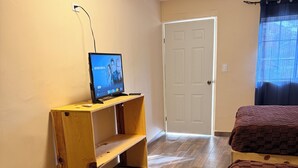 Une pièce avec un bureau en bois, un lit recouvert d’une couverture marron, une porte blanche et une fenêtre avec un rideau.