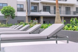 A row of grey lounge chairs with metal frames, positioned on a terrace with a view of a modern building and greenery. - Suite with Balcony at The Bantree (Accra)