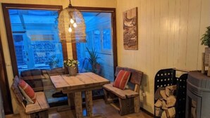 A cozy room with a wooden table, benches, a hanging light fixture, a fireplace, and a view through a large window. - Quiet vacation home "Zum Holzwurm" with large terrace & winter garden! (Molbergen)