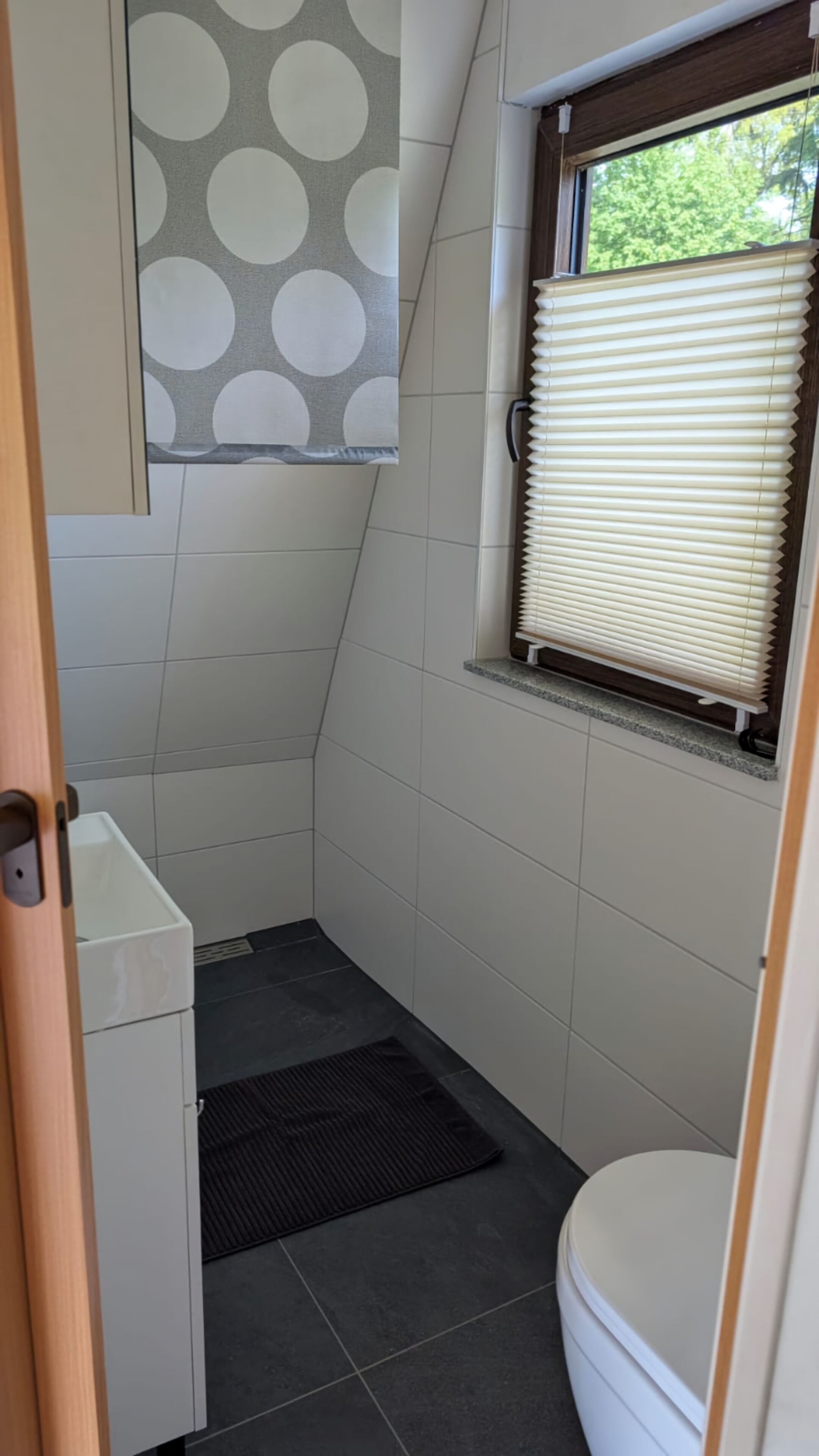 A small bathroom with a toilet, a window with blinds, a towel rack, and a shower area.