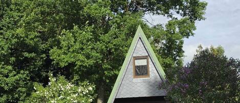 A small, triangular house with a sloped roof, surrounded by lush greenery and a bicycle parked outside.