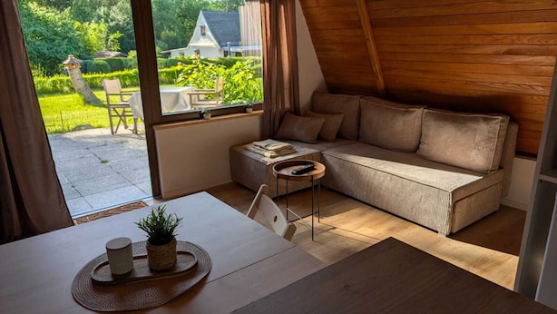 Ein gemütliches Zimmer mit Holzbalkendecke, einer Couch, einem kleinen Tisch mit Pflanze und Blick auf einen Garten und ein Haus.