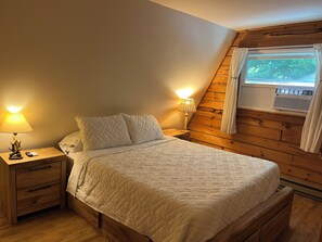 A bedroom with a wooden sloped ceiling, a bed with a quilted white duvet, two bedside lamps, and a nightstand with a drawer.