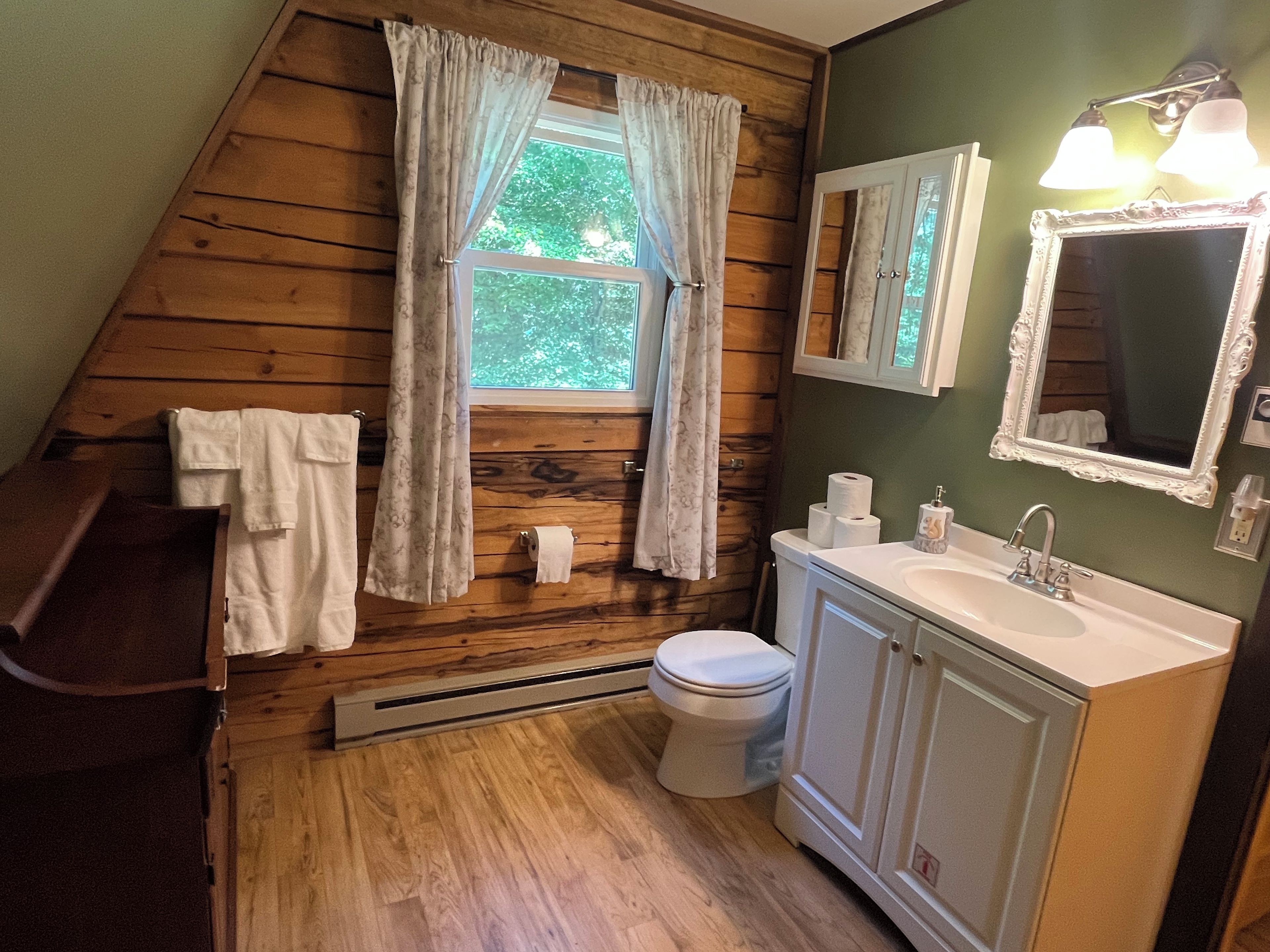 Une salle de bain avec des murs en bois, un lavabo blanc, des toilettes et une fenêtre avec des rideaux.
