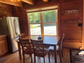 Ein Holzblockhaus-Interieur mit Esstisch, vier Stühlen, einem Kühlschrank und einem Fenster mit Blick auf eine Straße und Grünflächen.