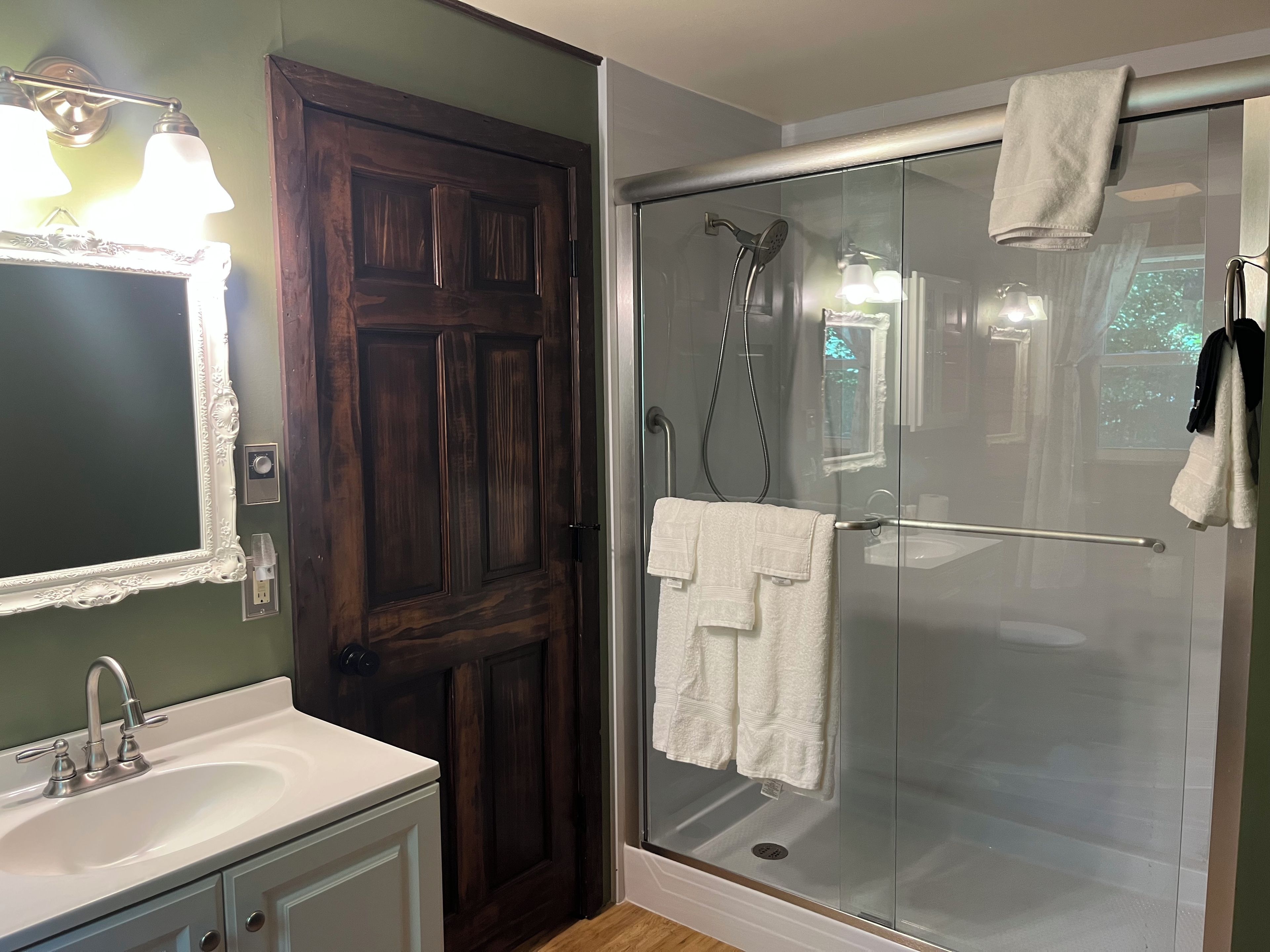 A bathroom with a wooden door, a glass shower enclosure, a sink, and a mirror with lights.