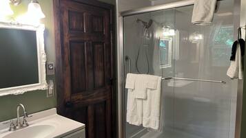 A bathroom with a wooden door, a glass shower enclosure, a sink, and a mirror with lights.