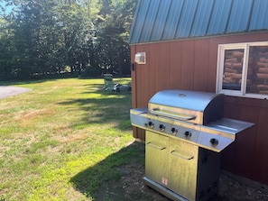 Ein tragbarer Grill mit vier Brennern, einem Wasserbehälter und einer Seitablage, neben einer Holzhütte mit Fenster.