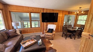 A wooden-paneled room with a sofa, a leather ottoman, a dining table with chairs, a flat-screen TV, and a chandelier.