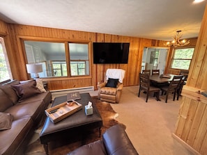 A wooden-paneled room with a sofa, a leather ottoman, a dining table with chairs, a flat-screen TV, and a chandelier. - RIVER SIDE walking distance to town/the lake & Rocky Mnt Natl Park! 3-bedroom (Grand Lake)