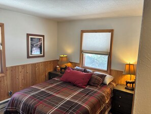 Ein Schlafzimmer mit kariertem Bettbezug, einem gerahmten Bild an der Wand, einem Fenster mit Jalousien und zwei Nachttischlampen.