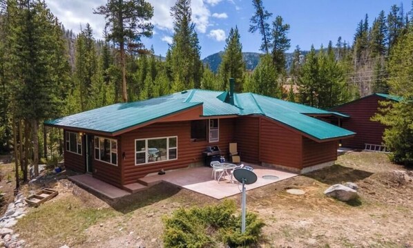 A cabin with a green roof, a patio with outdoor furniture, and a view of a forested mountain range. - RIVER SIDE walking distance to town/the lake & Rocky Mnt Natl Park! 3-bedroom (Grand Lake)