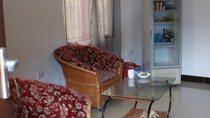 Uma sala de estar com uma mesa de centro de vidro, dois sofás estampados, uma geladeira repleta de bebidas e uma janela com cortinas.