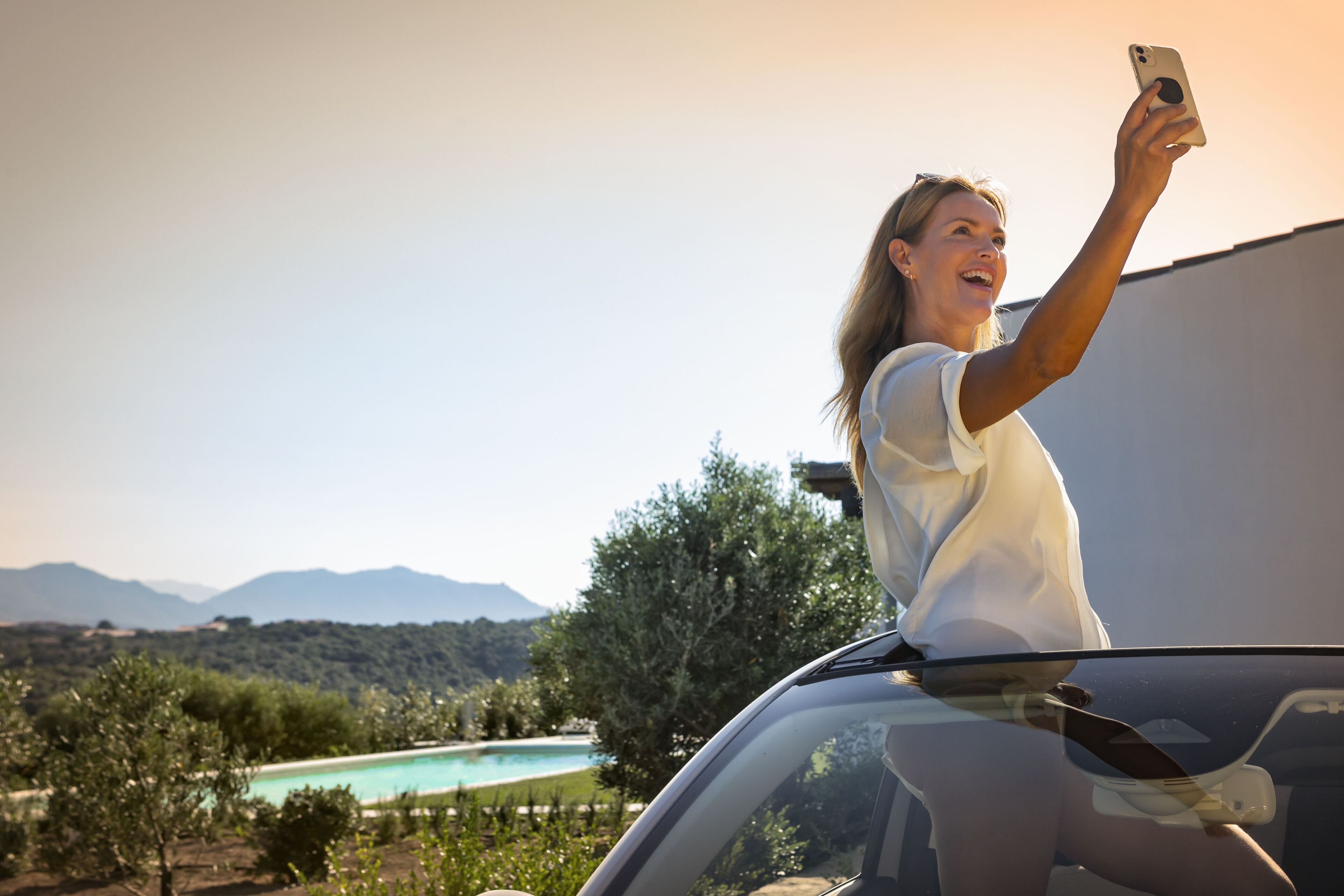 Una persona tomándose un selfie con un teléfono inteligente, de pie junto a un coche, con una piscina y montañas al fondo.