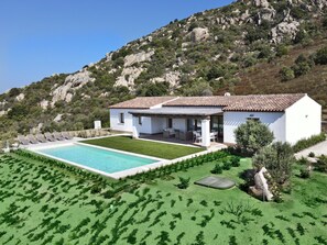 Eine weiße Villa mit Swimmingpool, Sitzgelegenheiten im Freien und einem Bergpanorama.