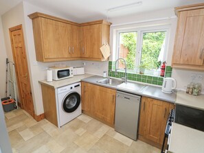 A compact kitchen with wooden cabinets, a washing machine, a dishwasher, a microwave, and a window with a view of greenery. - Tí Maolla (Hollymount)