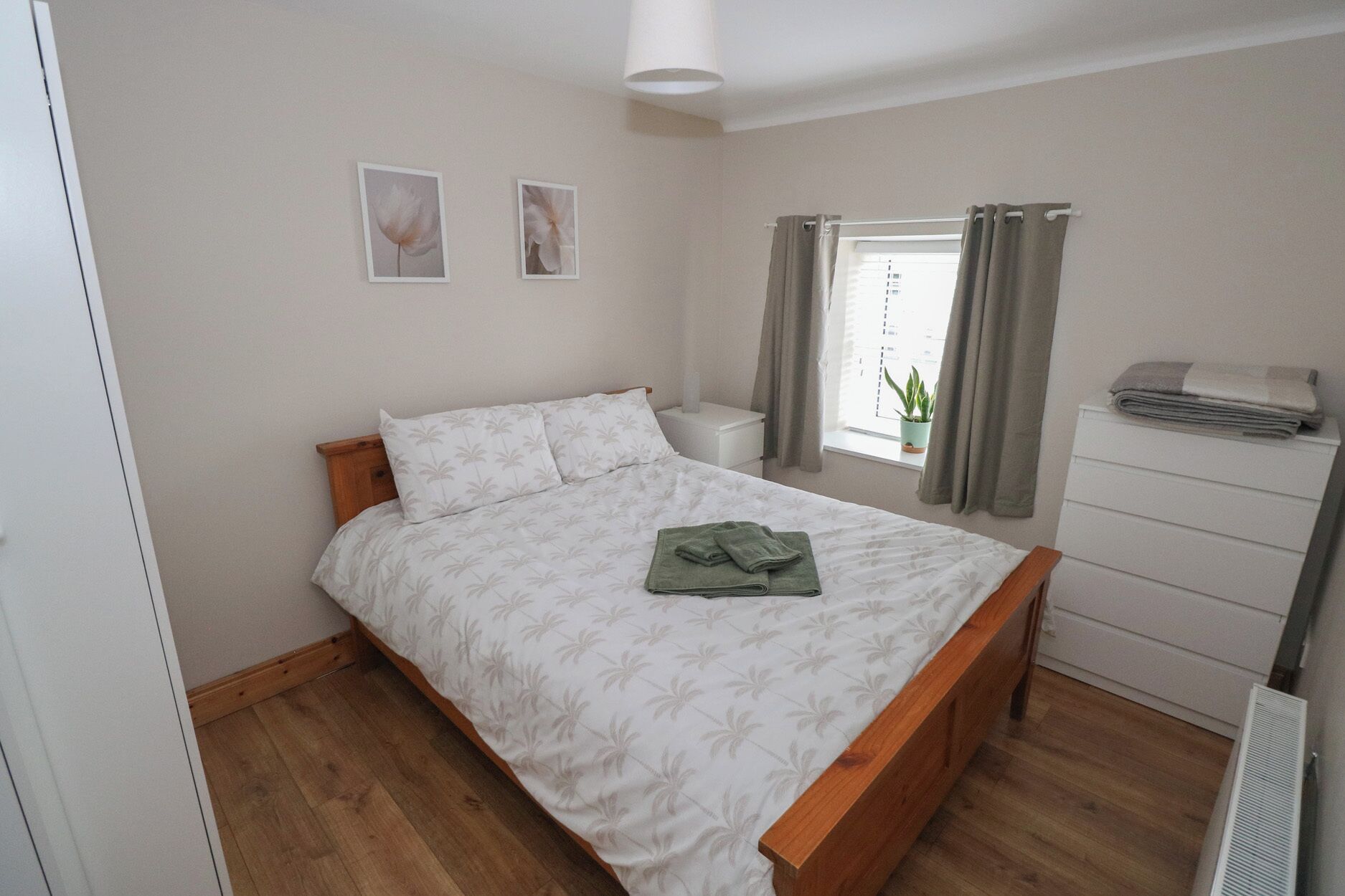 A bedroom with a bed, a nightstand, a window with curtains, a potted plant, and a dresser.