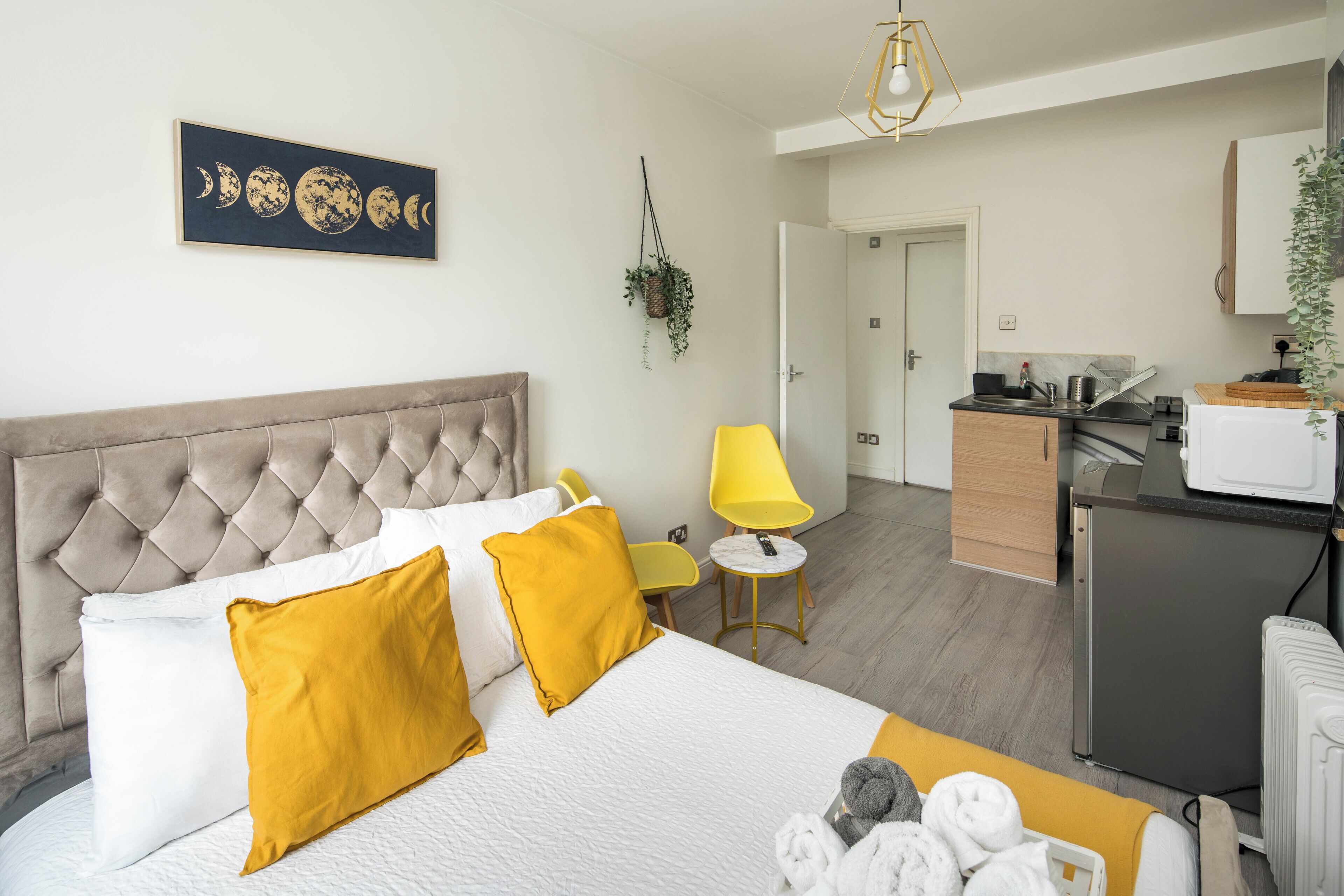 A hotel room with a bed, a small desk, a chair, a hanging plant, and a kitchen area.