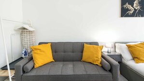 Un divano grigio con due cuscini gialli, un comodino bianco con una lampada e un letto grigio con biancheria da letto bianca.