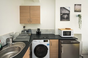 A compact kitchen with a washing machine, microwave, sink, and wooden cabinets. - Stylish London Stay | Jacuzzi, Sauna & Near the City Up to 6 People (London)