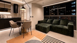A modern living room with a dark green sofa, a dining table with chairs, a kitchen area with a built-in oven, and a large window with city views. - Luxury Penthouse with Private Heated Pool & Christ the Redeemer Views in Ipanema (Rio de Janeiro)
