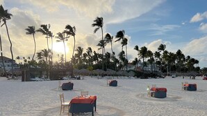 Een strand met ligbedden en tafels klaar voor dineren, omgeven door palmbomen en gebouwen op de achtergrond.