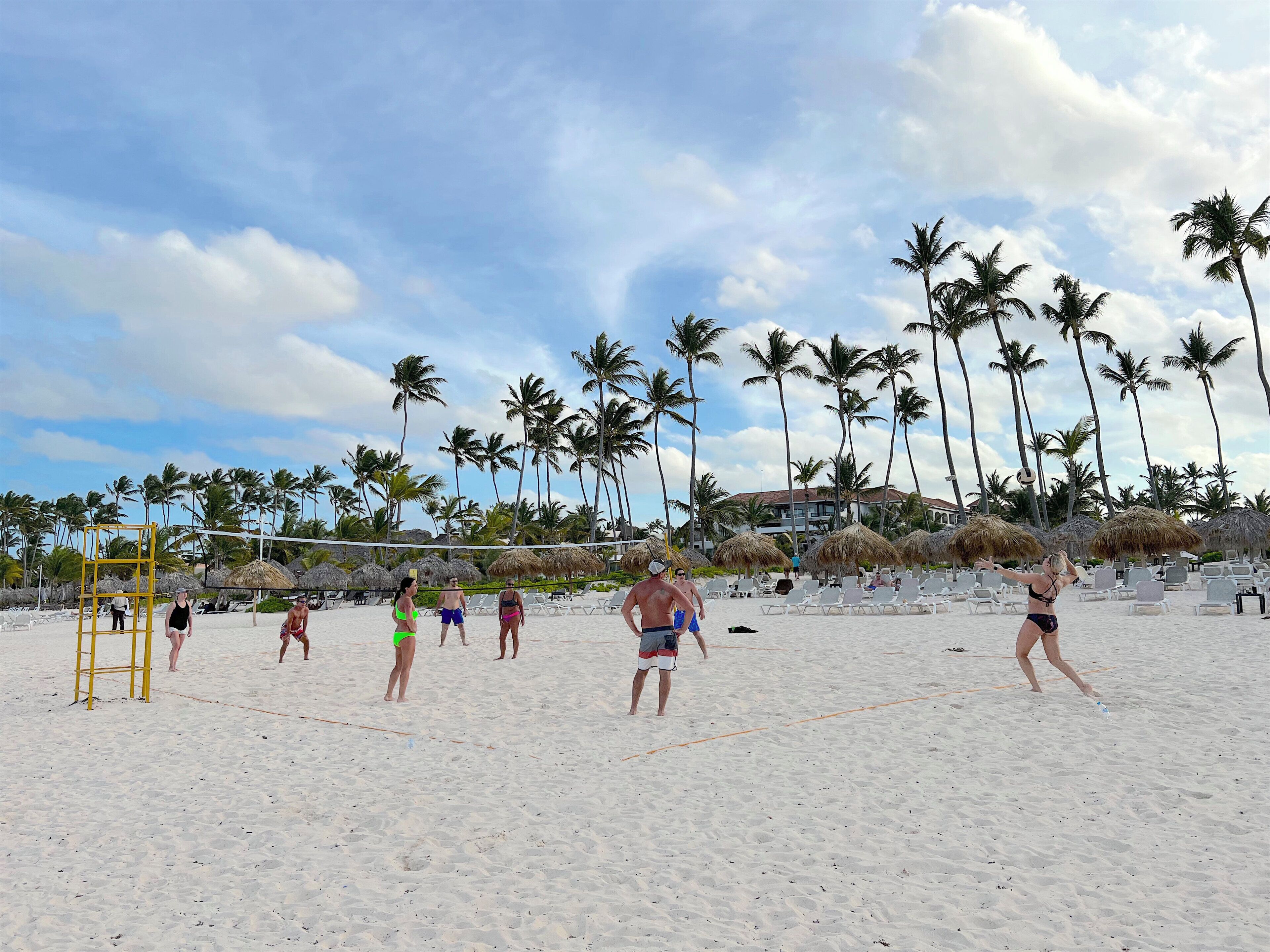 A beach volleyball game on a sandy court with palm trees and beach umbrellas in the background.