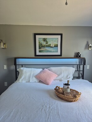 A neatly made bed with a white and pink pillow, a framed painting of a sailboat on a beach, and a wicker tray with a bottle and glasses on the bed.