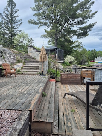 A wooden deck with a stone wall, a wooden chair, a gazebo, and a large tree.. Northern Paradise on the lake with water activities, Pet-friendly