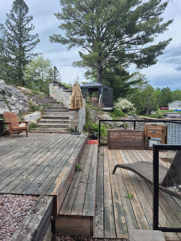 A wooden deck with a stone wall, a wooden chair, a gazebo, and a large tree.