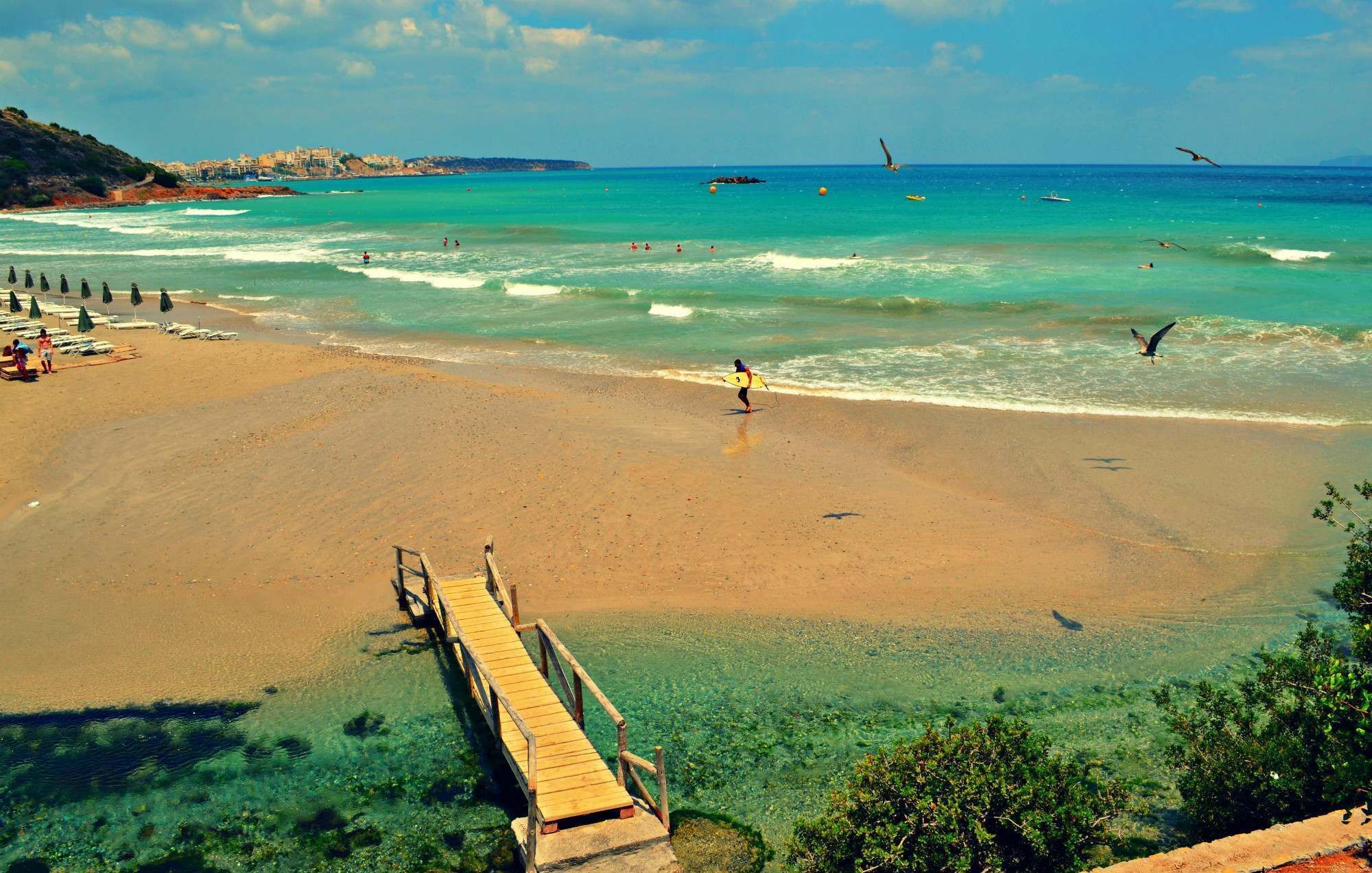 A beach with a wooden pier extending into the water, clear turquoise sea, and a distant shoreline with buildings.
