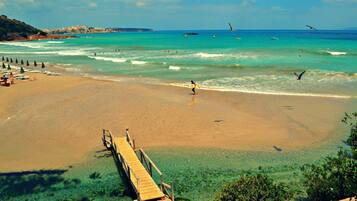 A beach with a wooden pier extending into the water, clear turquoise sea, and a distant shoreline with buildings.