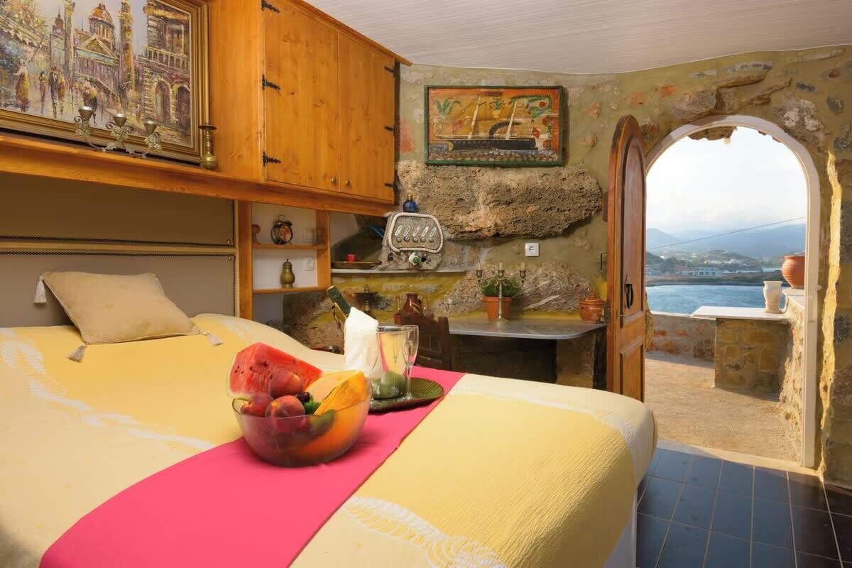 A bedroom with a bed, a fruit bowl, a table with a bottle, and a view of the sea through an archway.