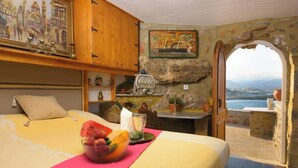 Ein Schlafzimmer mit einem Bett, einer Obstschale, einem Tisch mit einer Flasche und Meerblick durch einen Bogen.