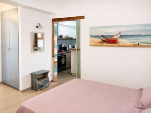 A bedroom with a bed, a small bedside table, a mirror, a painting of a boat on the wall, and a kitchen visible through an open door. - Votsalon Beach House - Family Retreat Zakynthos (Zakynthos)
