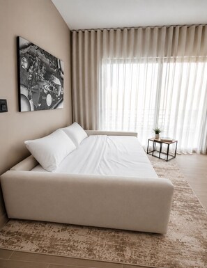 A modern bedroom with a large bed, a small side table, a potted plant, and a wall-mounted artwork.