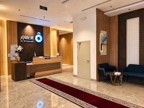 A hotel reception area with a wooden reception desk, a red carpet, and a blue sofa. - AlMohajer Serviced Apartments Airport  (jeddah)