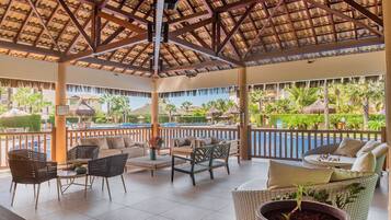 Un espacio lounge al aire libre con mobiliario de mimbre, vista a la piscina y un jardín tropical de fondo.