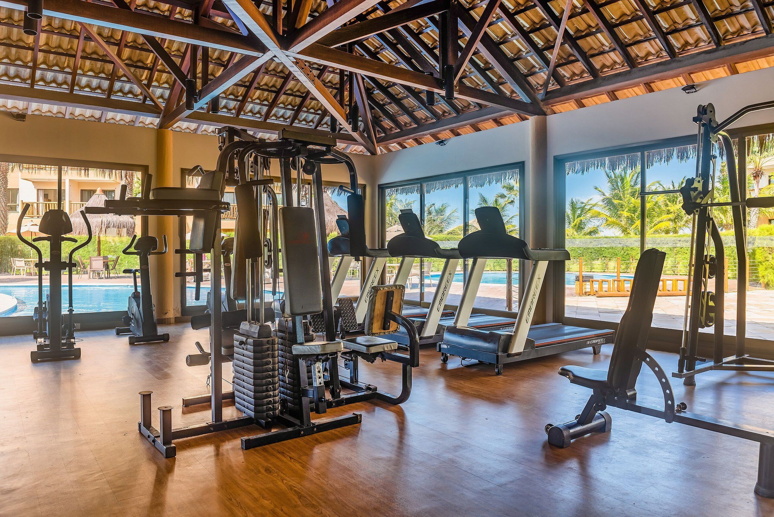 Un gimnasio con equipo de ejercicio, ventanas amplias y vistas a una piscina y palmeras.