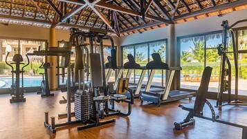 Un gimnasio con equipo de ejercicio, ventanas amplias y vistas a una piscina y palmeras.