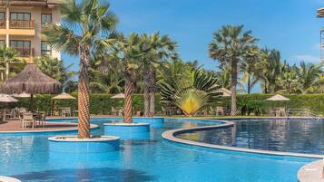 Un área de piscina en un complejo turístico con varias piscinas, palmeras y mobiliario exterior.