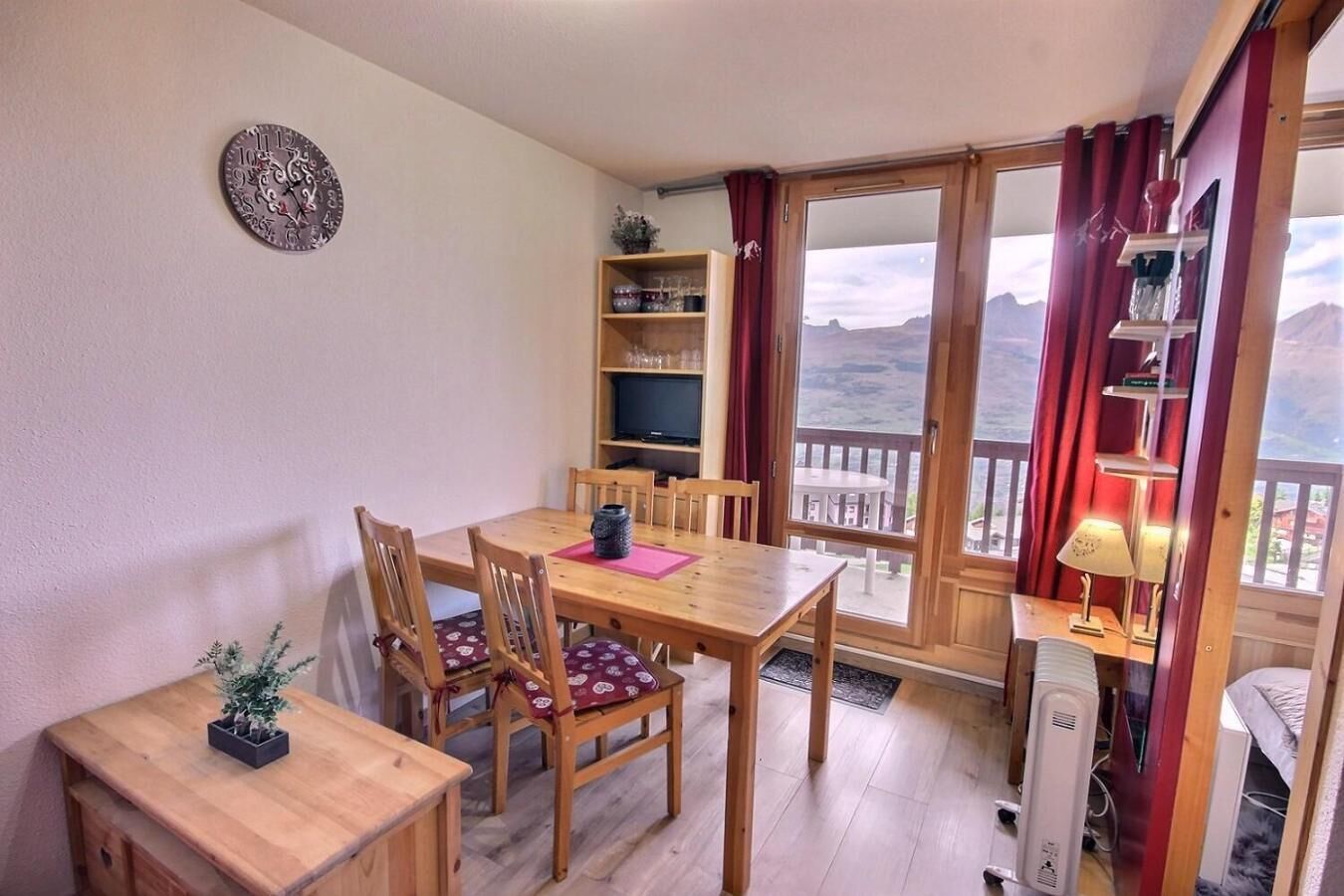 Een kamer met een houten eettafel, stoelen, een televisie op een plank, een balkon met uitzicht en een klok aan de muur.